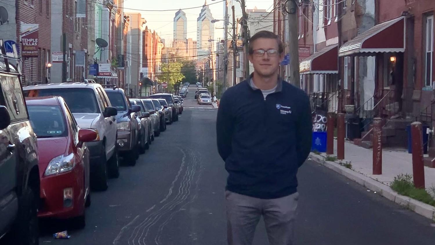 A man with light brown hair and glasses in a dark blue long sleeved shirt, gray pants and white sneakers is standing in the middle of the street with the Philadelphia city skyline in the background and parked cars on the left